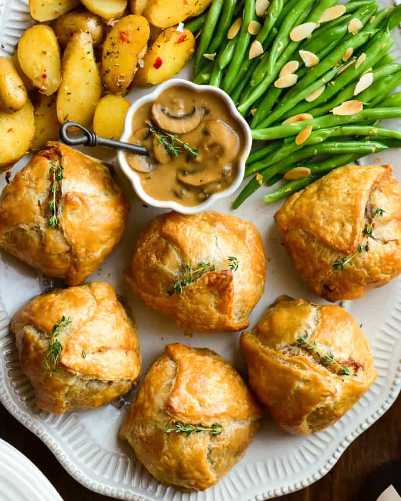 An oval platter with six individual ground beef wellingtons, a bowl of marsala mushroom sauce, potatoes and green beans.