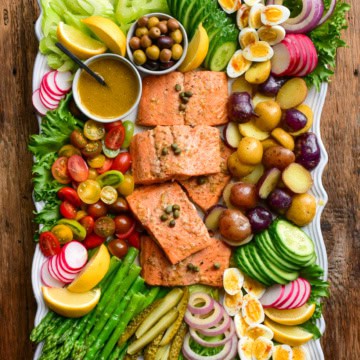 A large platter of a Niçoise Salad with Salmon. Five pieces of salmon are centered in the platter and are surrounded by various vegetables, eggs and pickles.