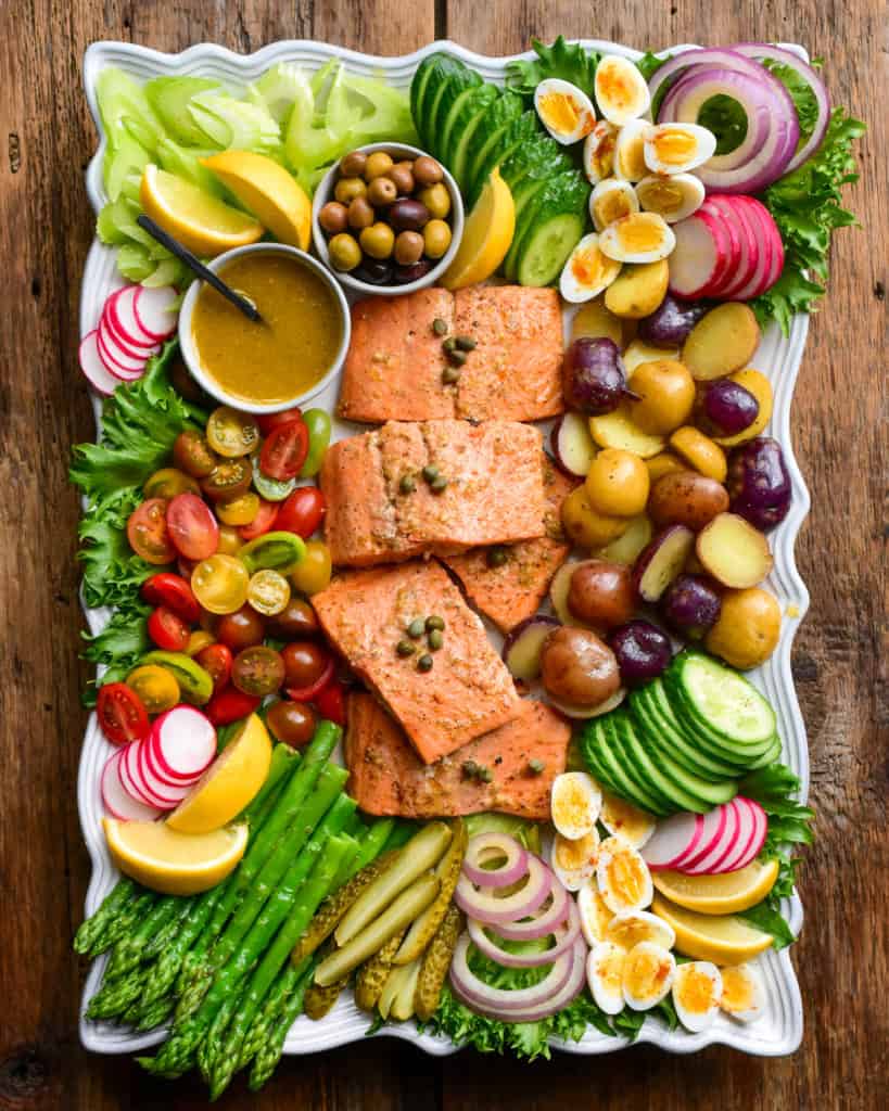 A large platter of a Niçoise Salad with Salmon. Five pieces of salmon are centered in the platter and are surrounded by various vegetables, eggs and pickles.