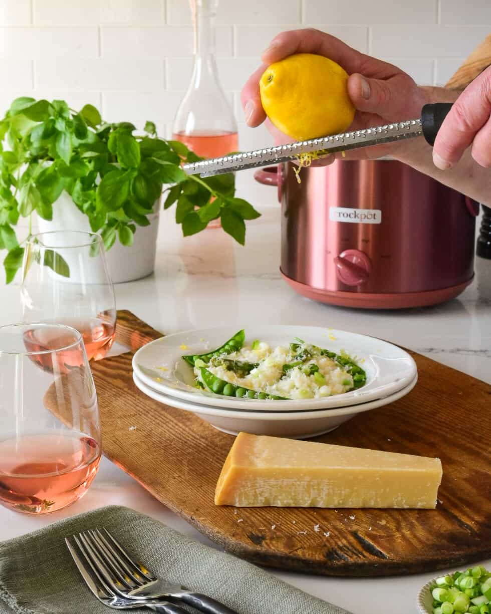 A plated bowl of a slow cooker spring risotto with asparagus and peas get lemon zested on it. With a rose copper Crockpot in the background.