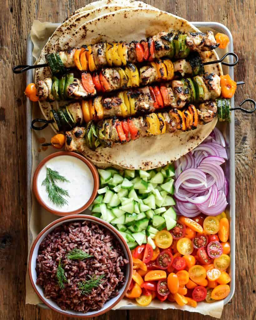 A pan with four chicken and vegetable skewers on top of a stack of large pitas. Served with rice, tzatziki, chopped cucumber, tomatoes and onions.
