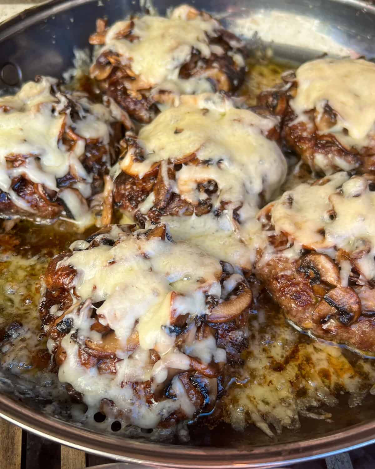 A skillet with six burger patties topped with mushrooms and melted cheese.