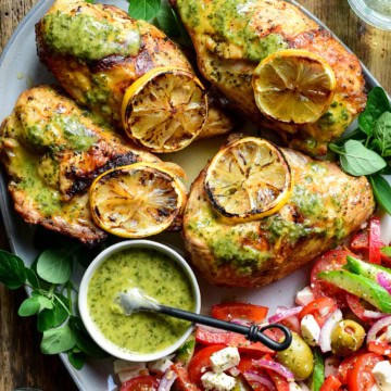 Four grilled and charred chicken breasts with charred lemon slices and served with a tomato salad.