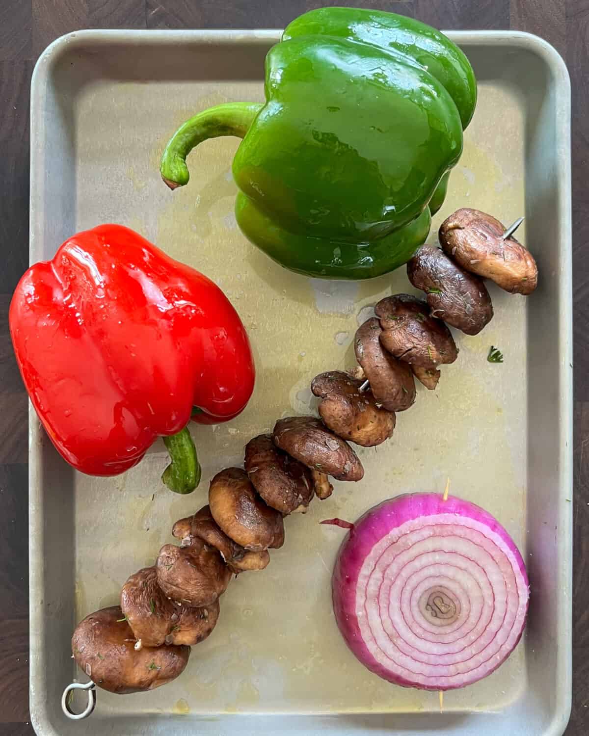 An oiled red and green pepper, a slice of red onion and a skewer of mushrooms are ready for the grill.