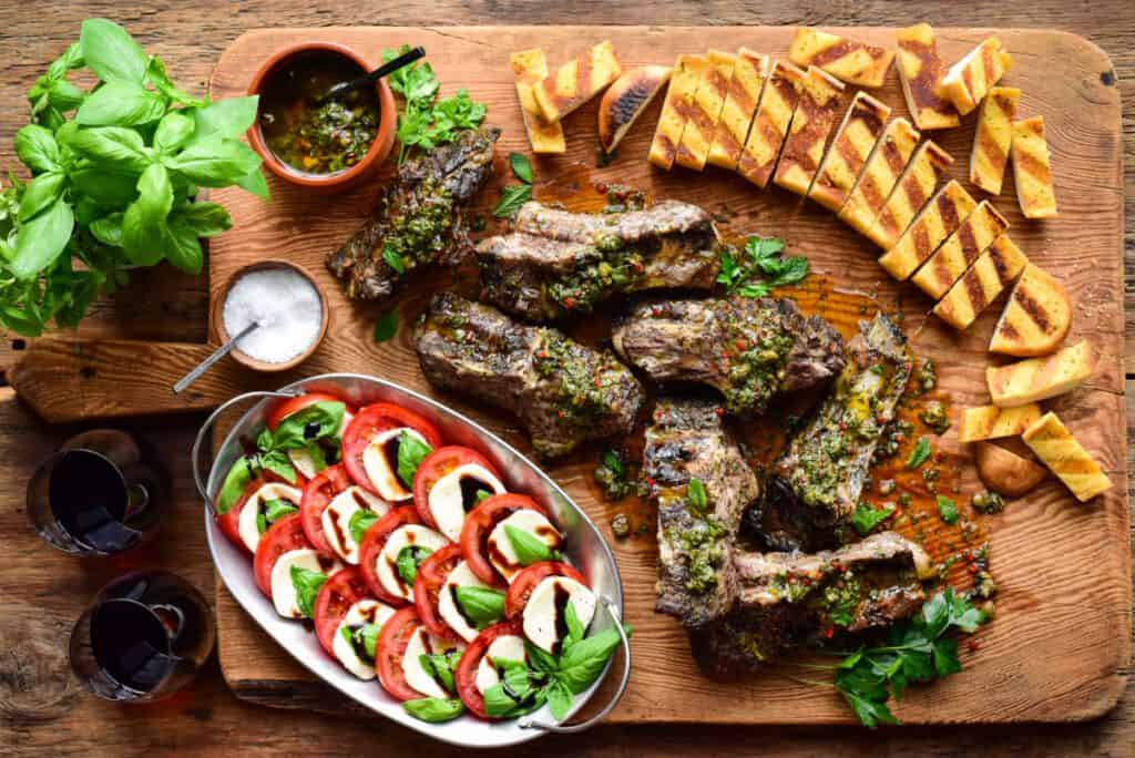 Seven grilled big ribs with a green salsa on a board with grilled garlic toast and a Caprese salad.