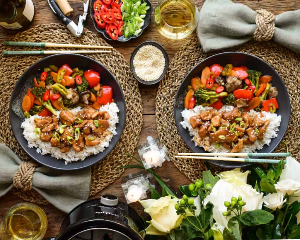 A place setting for two of a chicken teriyaki meal with chopsticks. Also on the table are candles, flowers, a crockpot, wine bottle, wine glasses, an opener and a slow cooker.