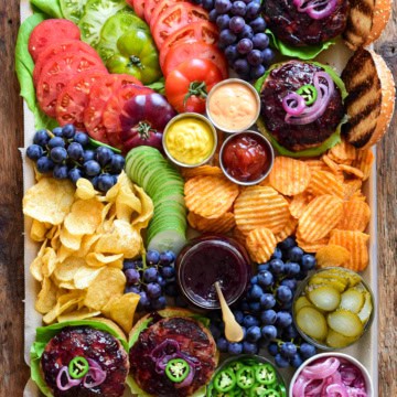 A top down image of a colourful tray with burgers, grape jam, blue grapes, tomatoes, chips and condiments.