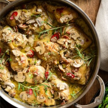 A close up of a finished chicken coq au vin blanc dish. The chicken is in a mushroom sauce with herbs and red peppers.