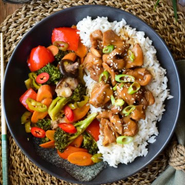 A black plate with rice and chicken on one side and bright cooked vegetables on the other side.