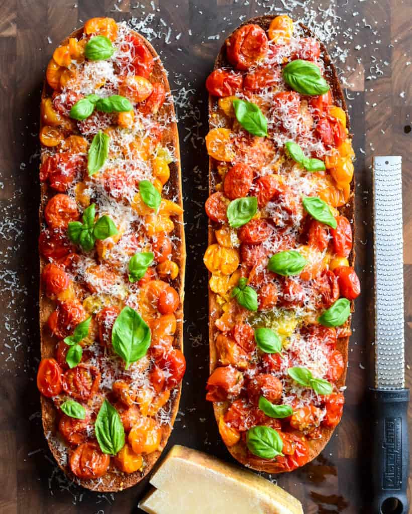 Two large half loaves of toast with tomato confit topped with grated parmesan and basil. A block of cheese and grater are beside the toast.