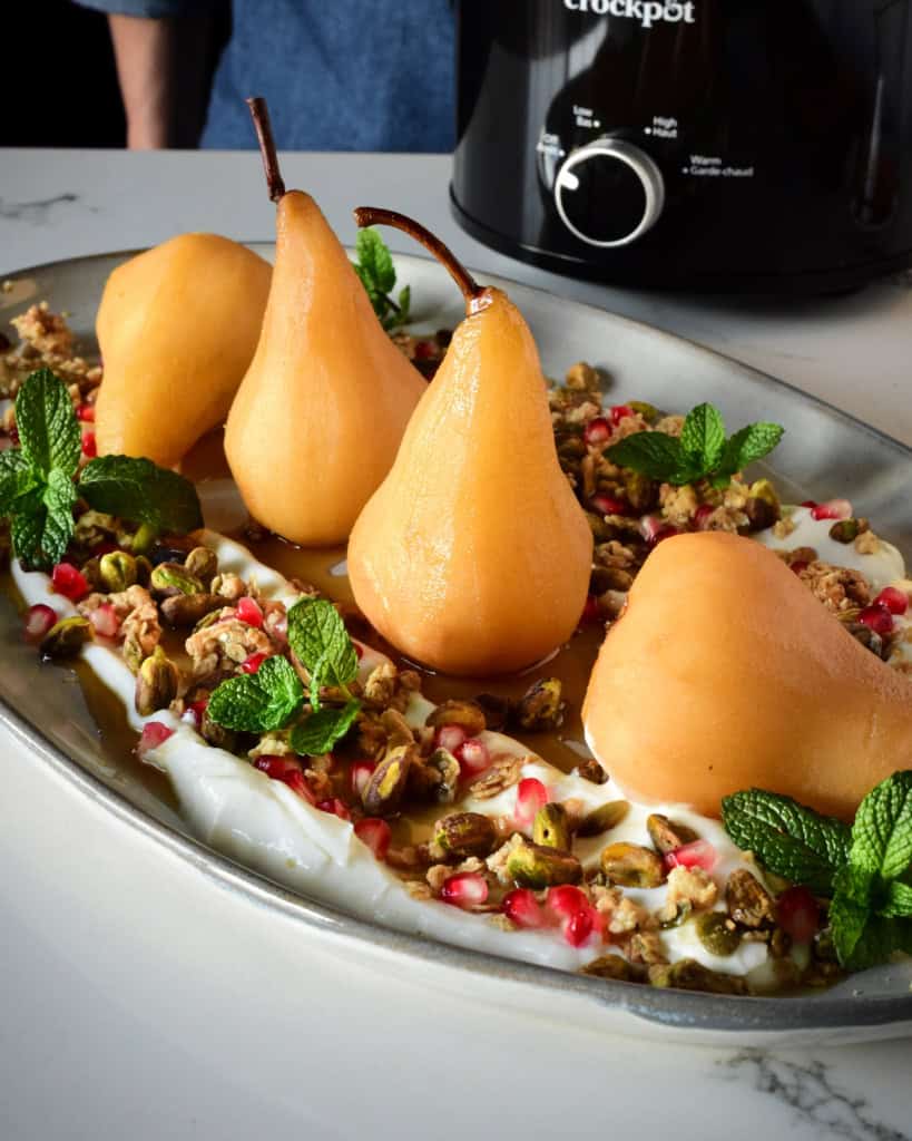 Four poached pears lined up on a platter surrounded with yogurt, nuts and granola with a black crockpot in the background.