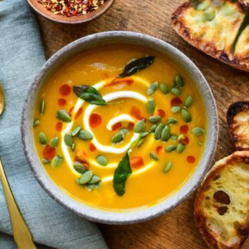 A bowl of Honeynut Squash Soup topped with a swirl of Greek yogurt, sprinkled with pepitas, chili oil and fried sage. Extra topping and three slices of baguette toast surround the bowl.