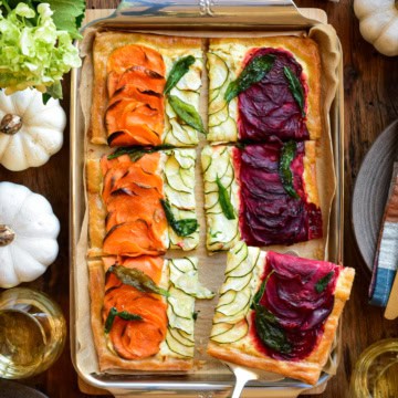 A puff pastry with three rows of vegetables, sweet potato and beet on the outside and zucchini on the inside. The tart is cut into six pieces. The table setting is a fall theme.