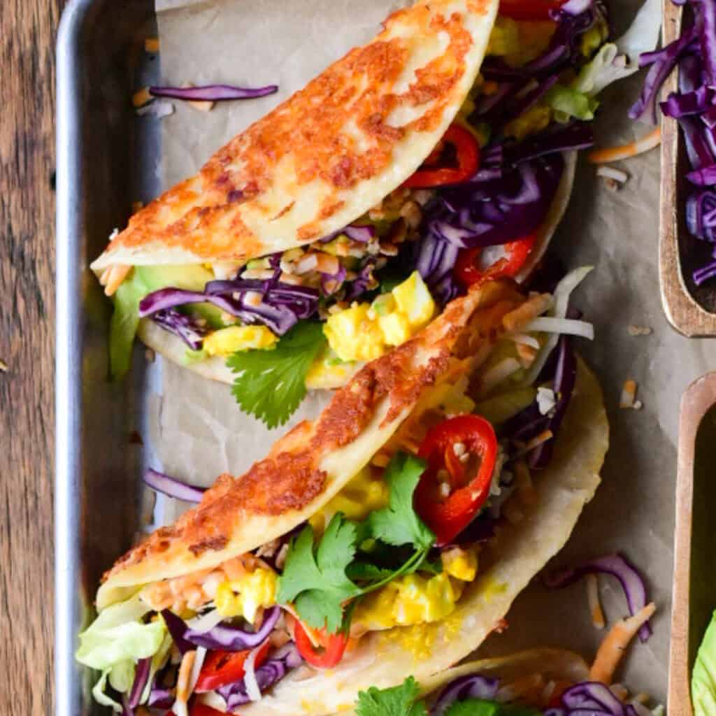 A close up of a cheesy tortilla taco with egg salad and other colourful toppings.