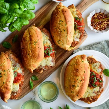 Three crispy chicken sandwiches that look like chicken parmesan sandwiches on a tray, and one is on a plate. Served with extra sauce, basil, and drinks.