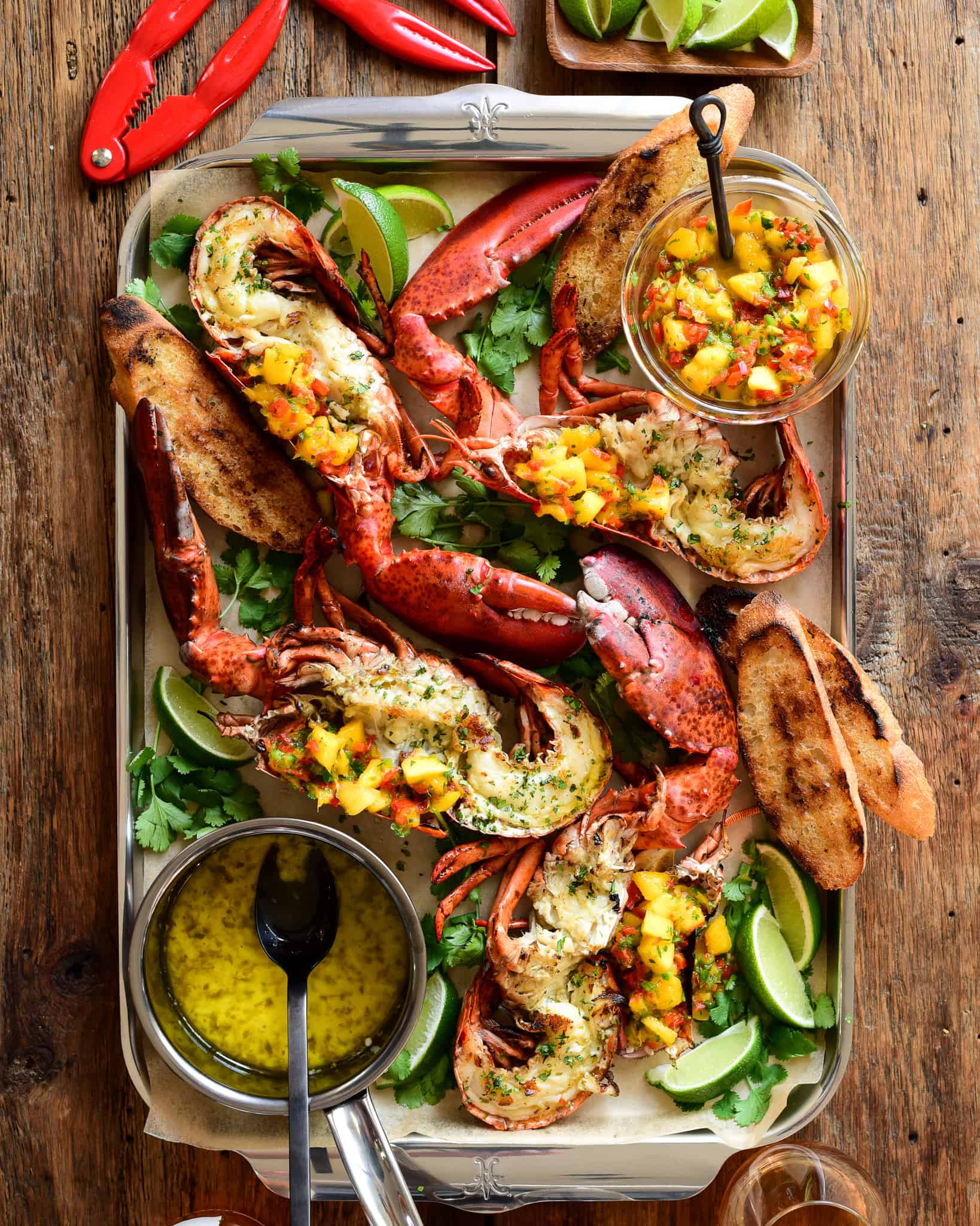 Grilled half lobsters topped with a Mango-Jalapeño Salsa on a tray with a pot of garlic-chive butter.