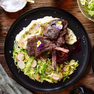 Two venison chops are leaning over mashed potatoes, a brussels sprout slaw alongside and Jalapeño Sauce on a black plate.