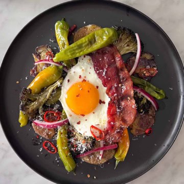 A plate of Parmesan Smashed Potatoes and Eggs with roasted broccoli, shishitos, long red chilies, a crispy slice of bacon and marinated red onions.