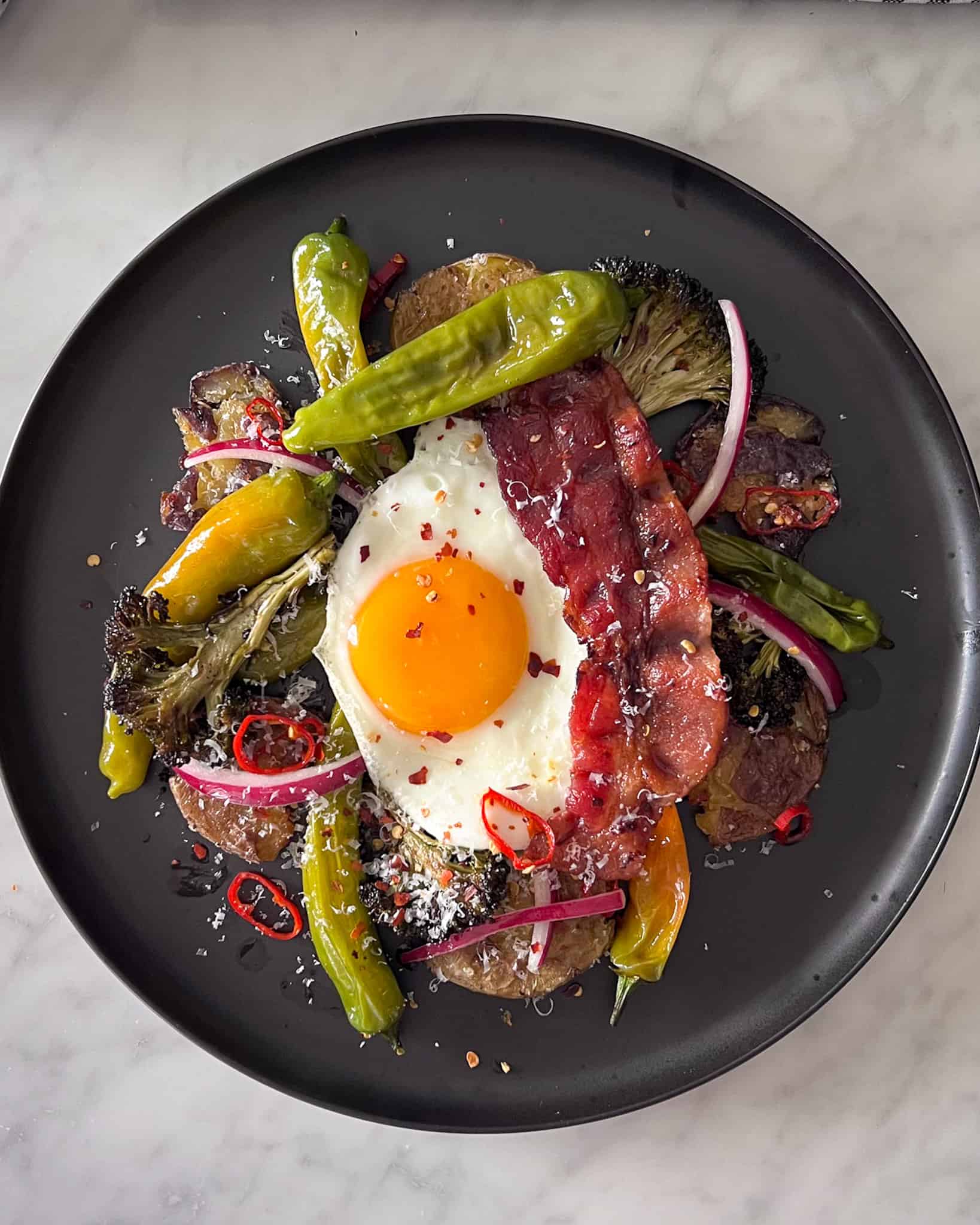 A plate of Parmesan Smashed Potatoes and Eggs with roasted broccoli, shishitos, long red chilies, a crispy slice of bacon and marinated red onions.