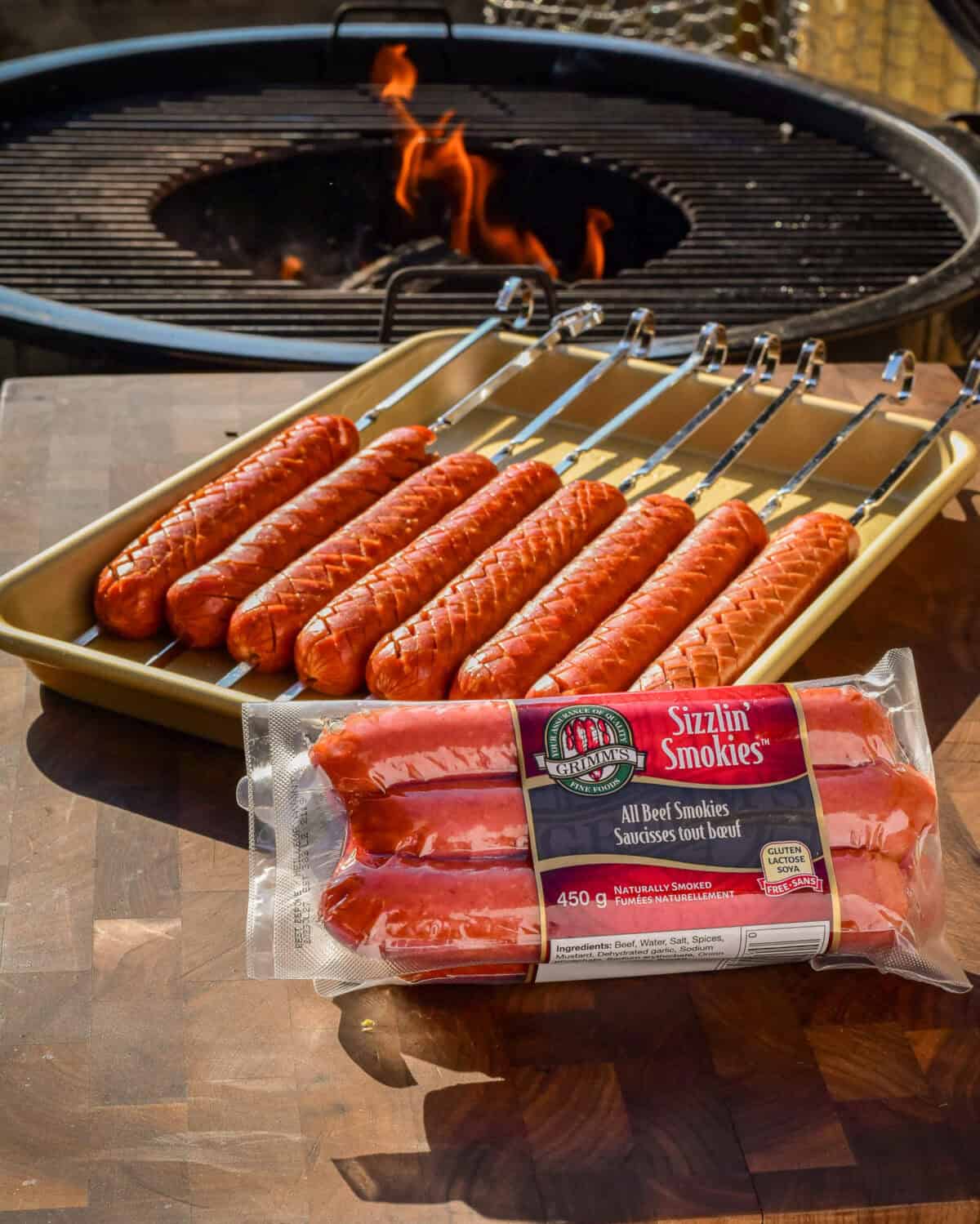 Crosshatched beef smokies on a tray beside a flaming BBQ ready to be grilled.