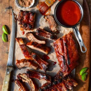 A cutting board with sauced baby back ribs and a pot of tequila BBQ sauce. A large knife has sliced most of the ribs.
