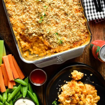 A tray of Buffalo Cauliflower Mac and Cheese with a portion on a plate. Served with crudités and blue cheese dip.