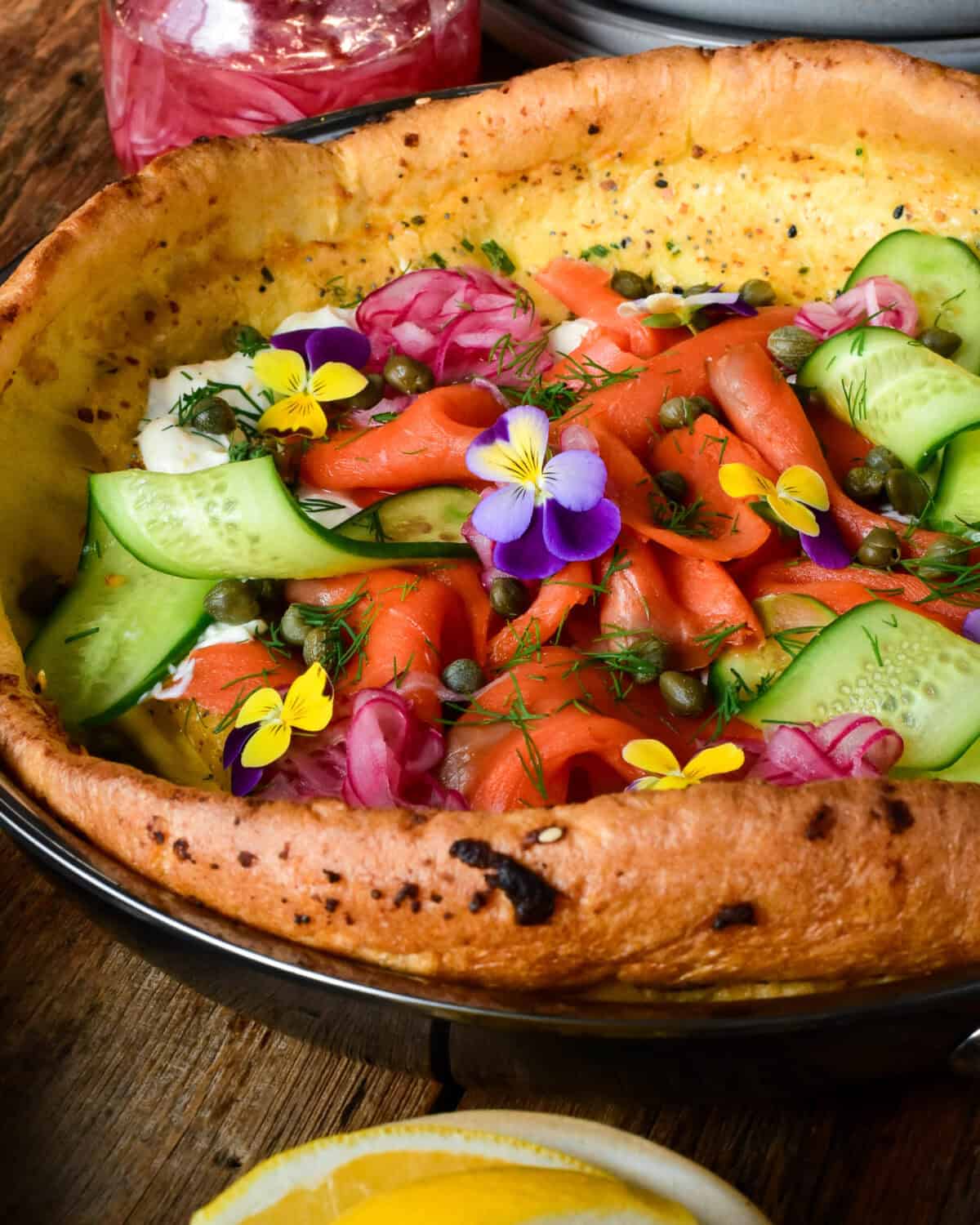 A close up look at a Dutch baby with Smoked Salmon, cream cheese, capers, pickled onions, cucumber, fresh herbs, everything bagel seasoning and edible herbs