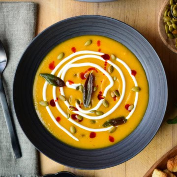 A bowl of Butternut Squash and Ginger Soup garnished with Greek yogurt, pepitas, chili oil, fried sage. Surrounded by toppings and croutons.