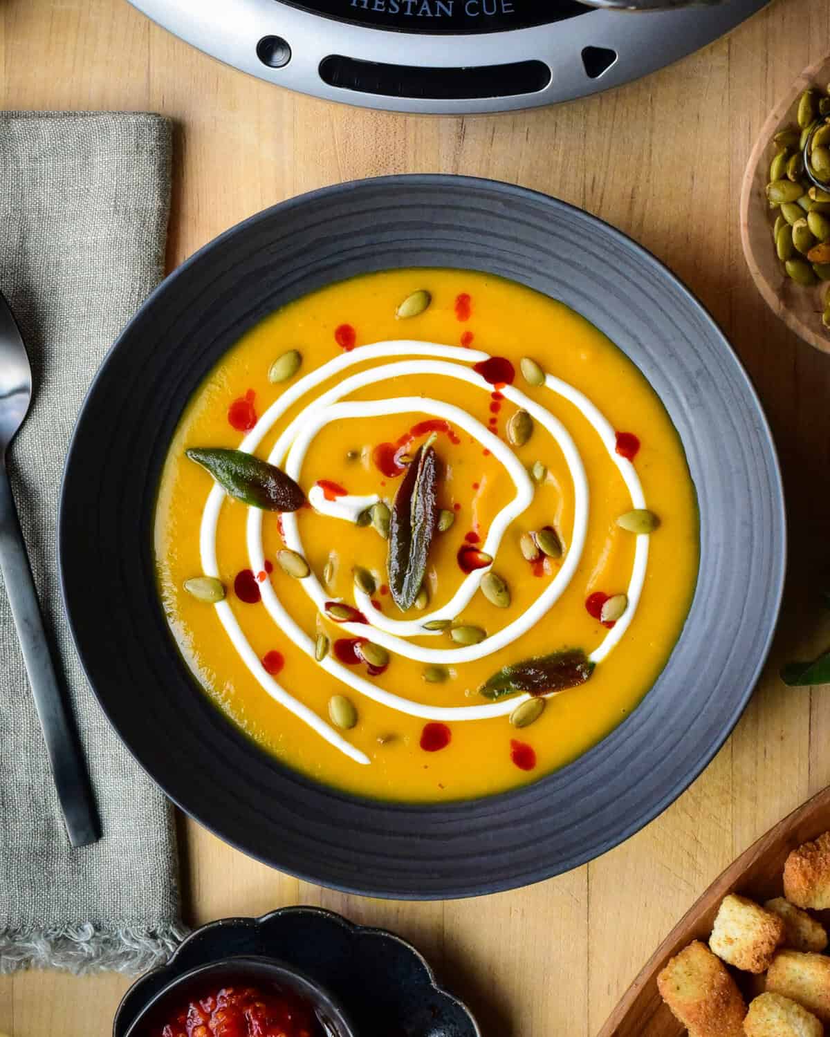 A bowl of Butternut Squash and Ginger Soup garnished with Greek yogurt, pepitas, chili oil, fried sage. Surrounded by toppings and croutons.
