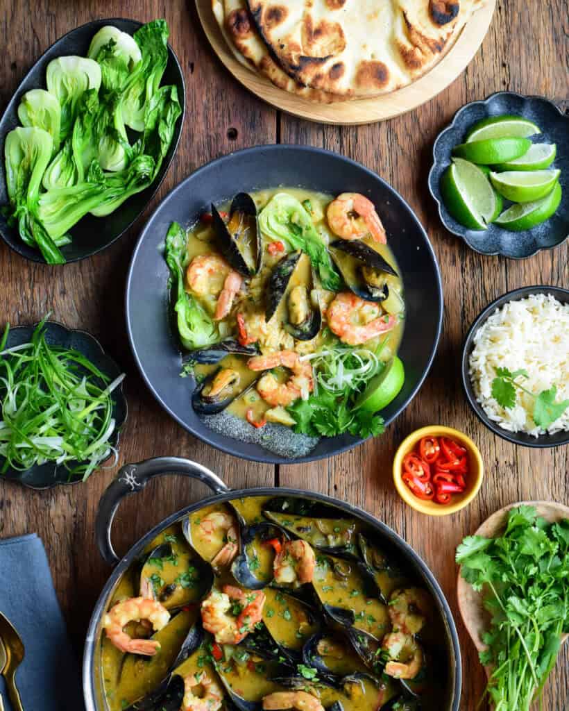 A bowl and pan filled with yellow curry and shrimp surrounded with garnishes.