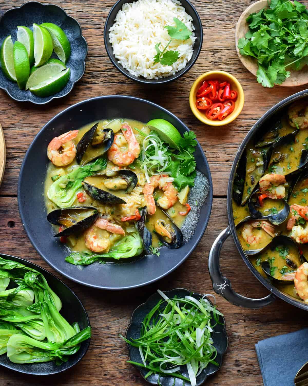 A bowl of yellow curry with shrimp and mussels, served with rice, green onions, cilantro, a lime wedge and served with warm naan.