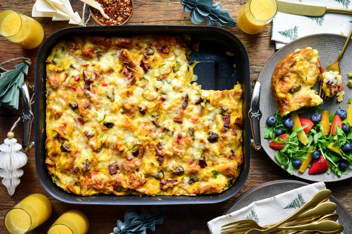A roasting pan with a make ahead croissant breakfast casserole, with a piece taken out and served with fruit.
