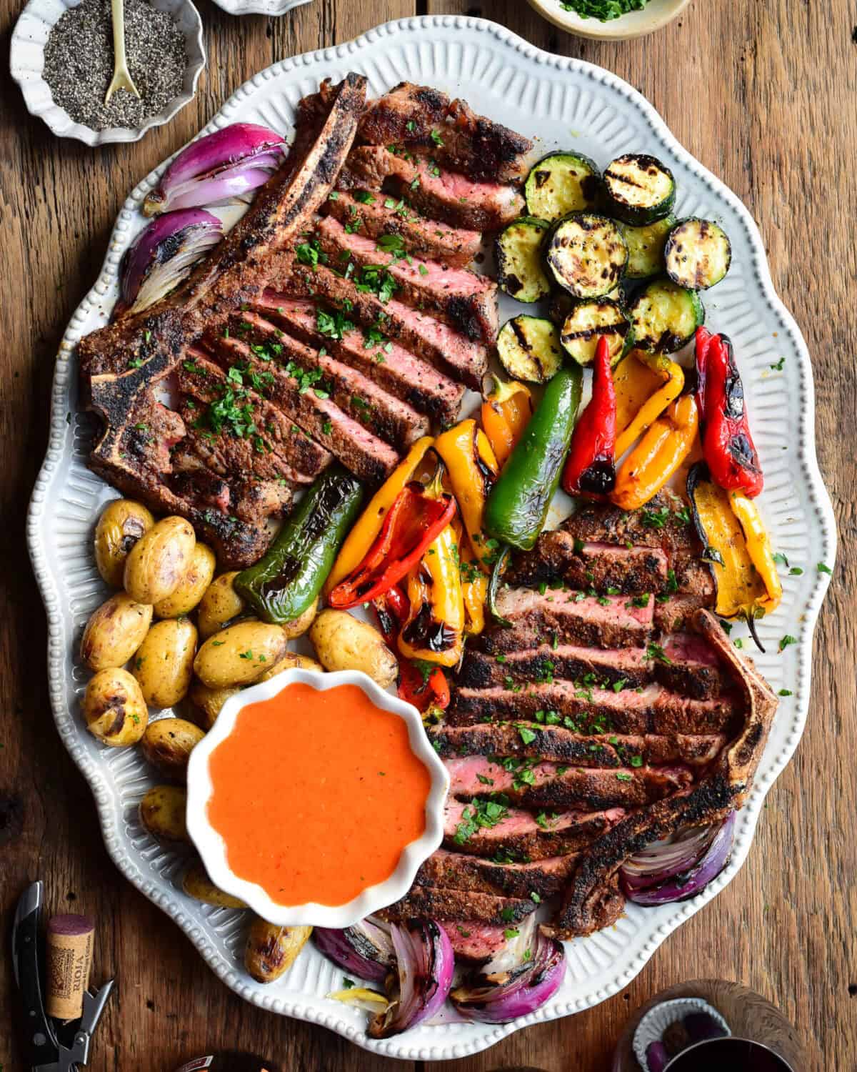 Two sliced Spanish Style Steaks with assorted grilled vegetables and a bowl of Spanish steak sauce.