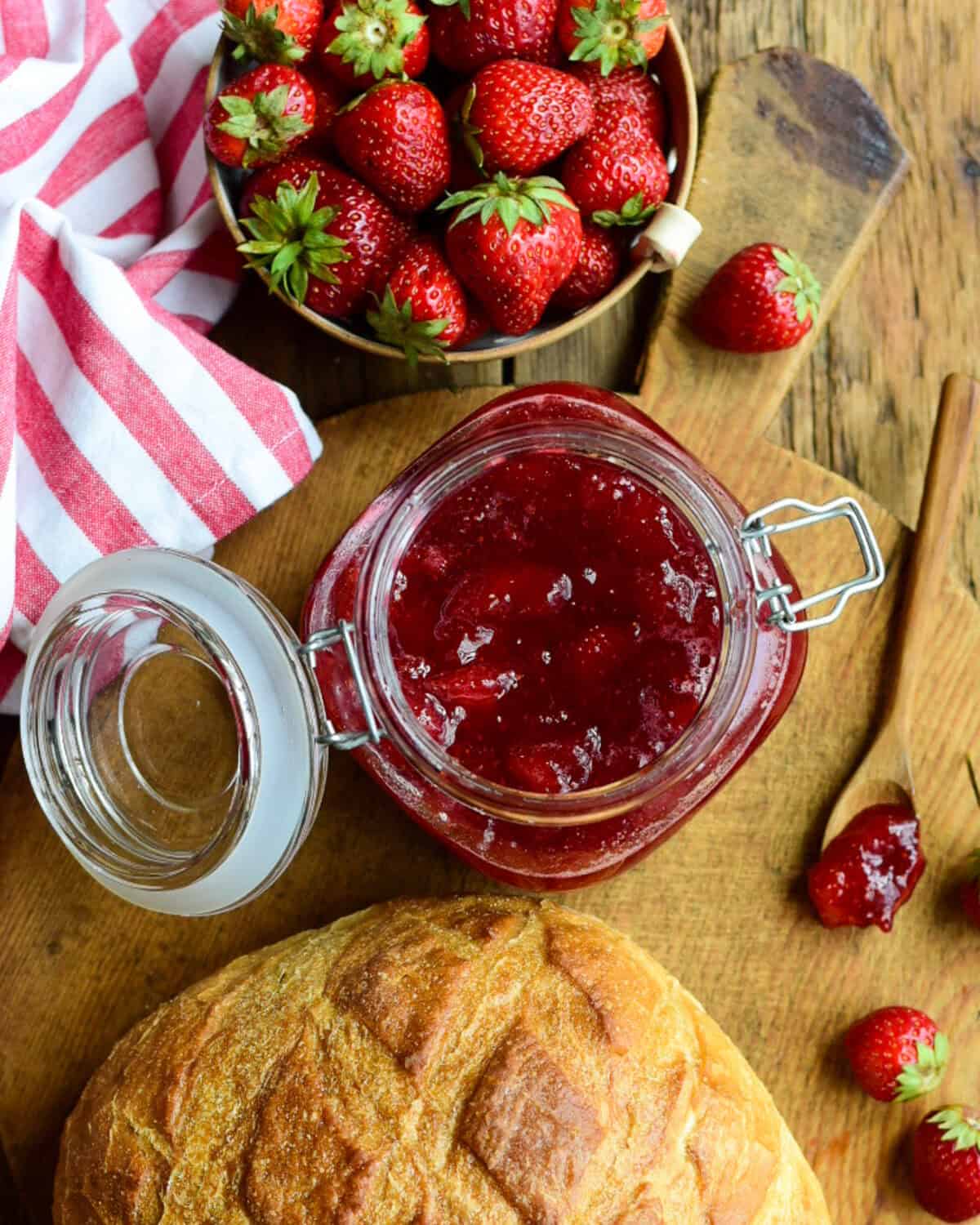 A large mason jar filled with Strawberry-Ginger Jam on a board with a sourdough loaf and fresh strawberries.