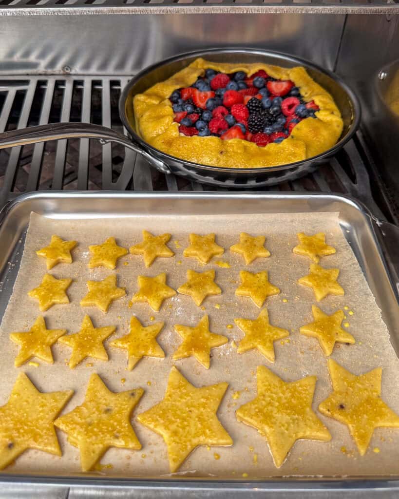 A skillet with a berry galette and a sheet pan with star 'cookies' both on the grill.