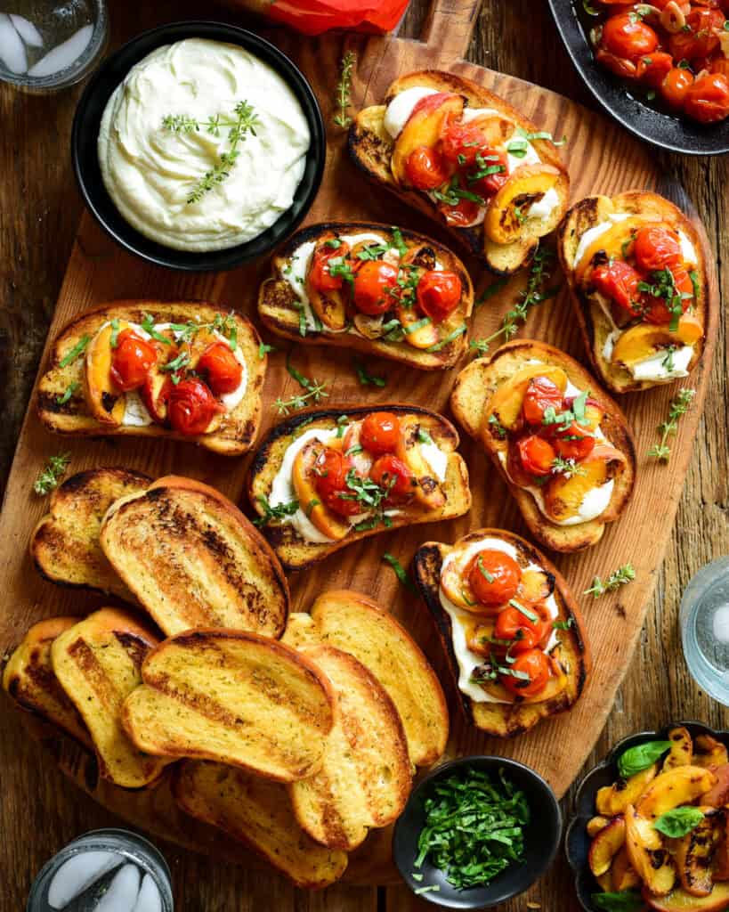 A wooden board with Grilled Peach & Tomato Crostini with Whipped Feta appetizers. Served with extra toppings and grilled garlic bread.