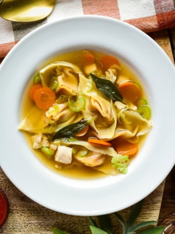 Bowl of autumn honeynut squash wonton soup featuring wontons, carrots, celery, and green onions in a clear broth, served in a white bowl on a rustic wooden table.