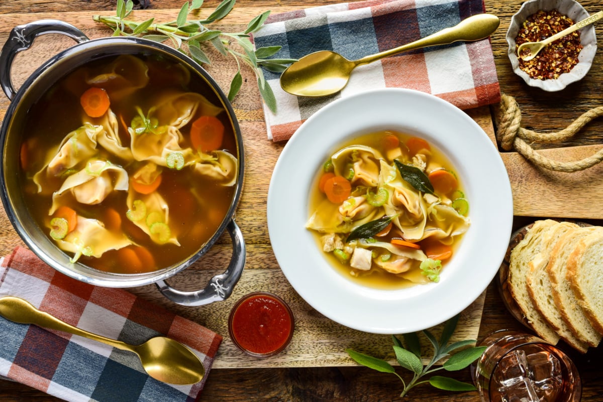 A bowl of warm wonton soup with carrots, greens, and broth, served with bread slices and spices on a rustic wooden table.