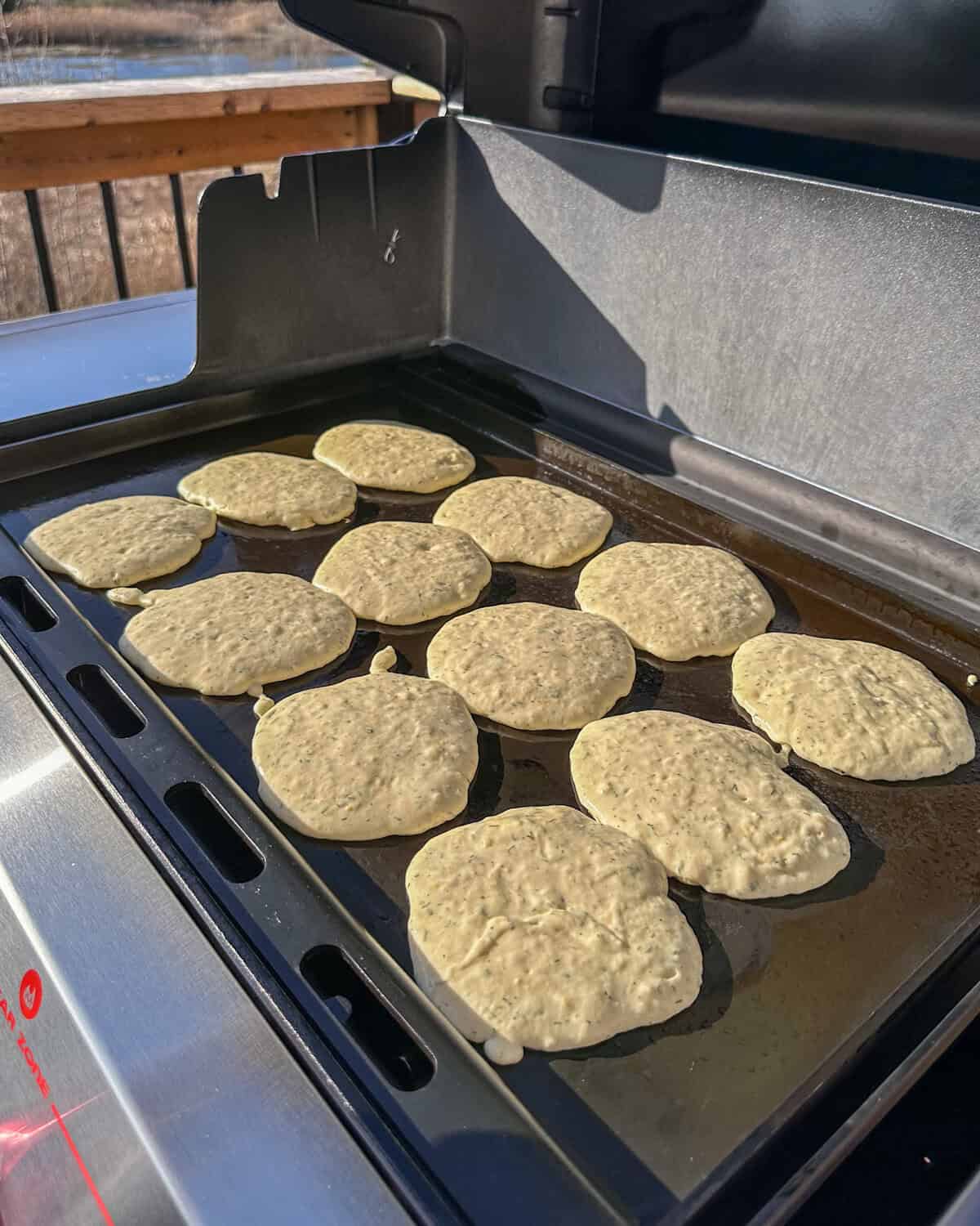 Twelve pancakes on a grill forming bubbles.