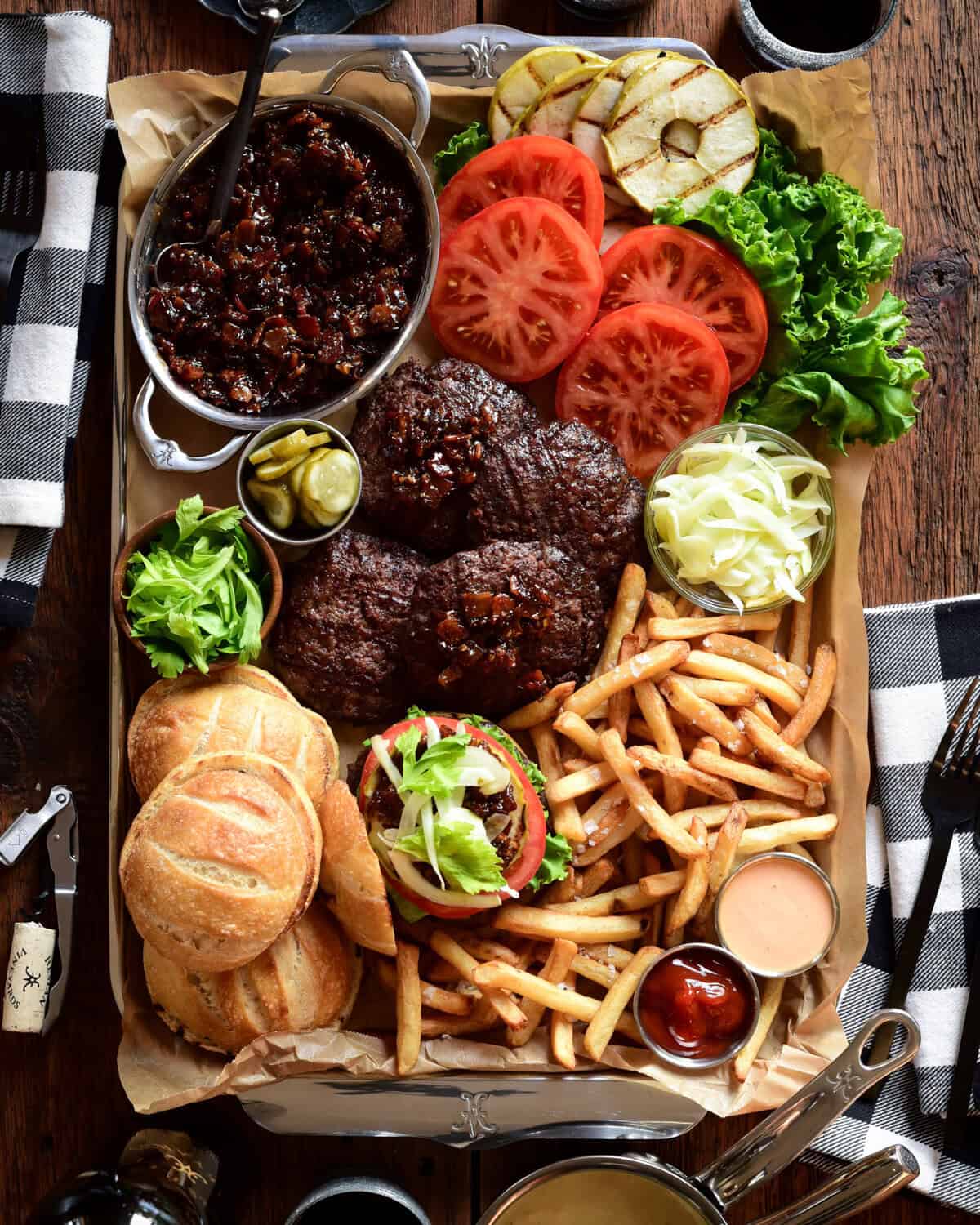 A serving tray with Espresso Bacon Jam Burgers with toppings, fries and a pot of smoked Gouda Sauce.