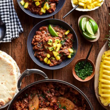 Two bowls and a pot of Caribbean Baked Beans with Jerk-Spiced Pork Shoulder. Served with pineapple, naan and lime wedges.