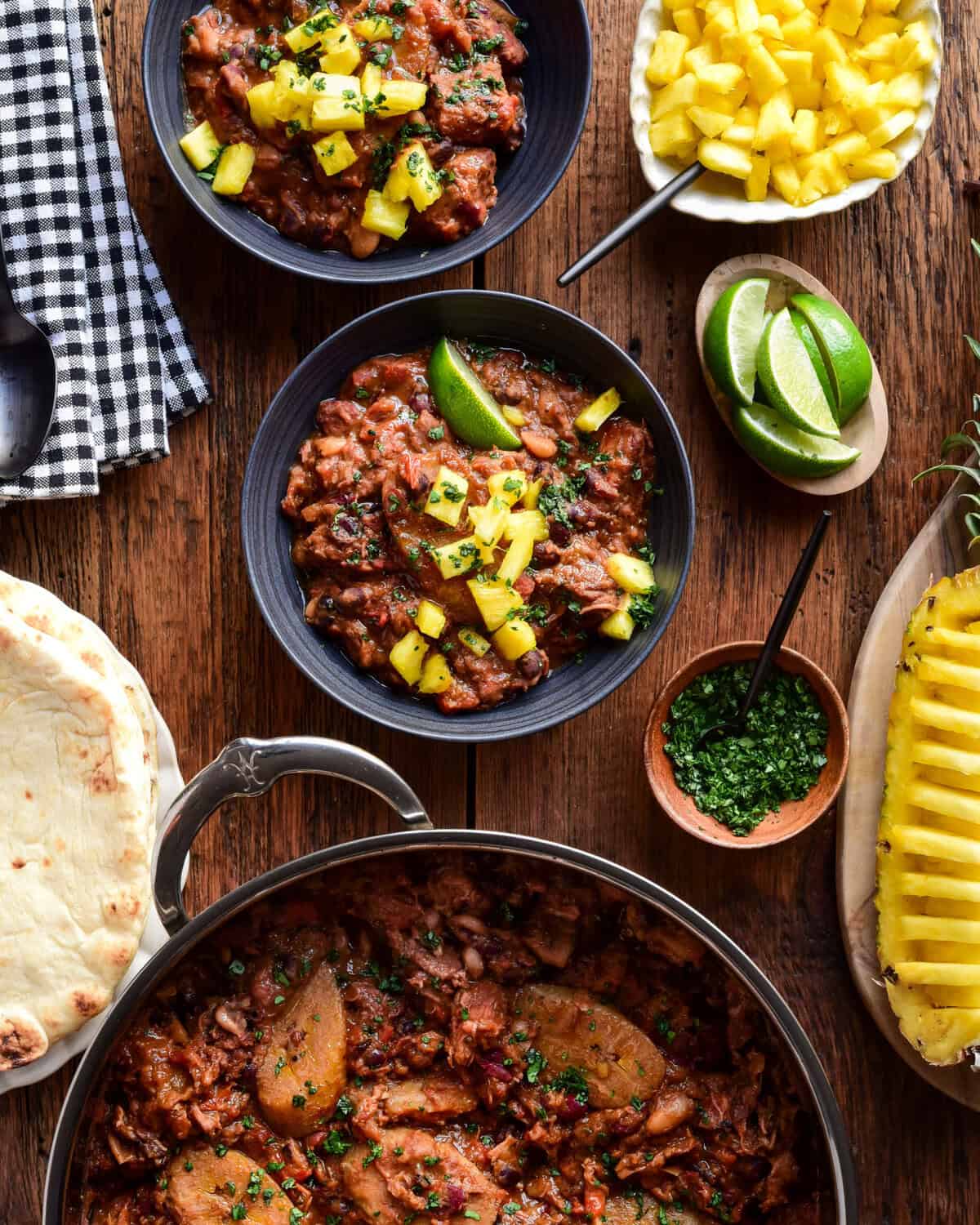 Two bowls and a pot of Caribbean Baked Beans with Jerk-Spiced Pork Shoulder. Served with pineapple, naan and lime wedges.