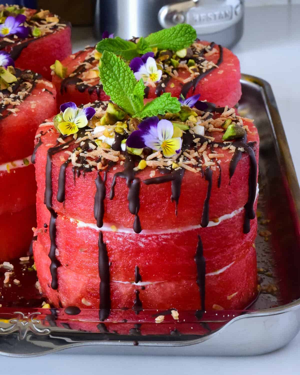 A close-up of mini watermelon cakes with coconut whipped cream, dark chocolate drizzle and topped with pistachios, toasted coconut, mint and edible flowers.