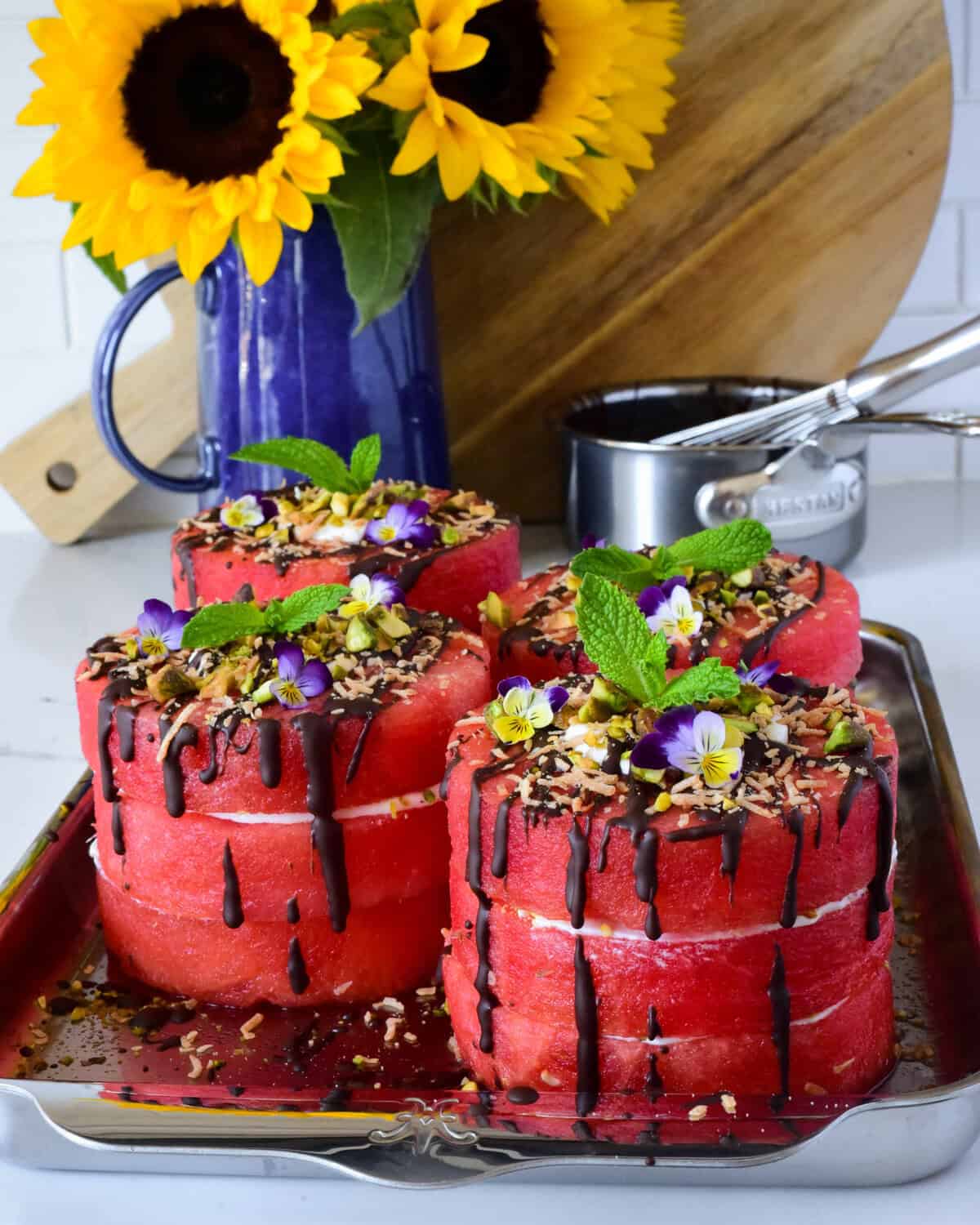 Four mini watermelon cakes with coconut whipped cream, dark chocolate drizzle and topped with pistachios, toasted coconut, mint and edible flowers.