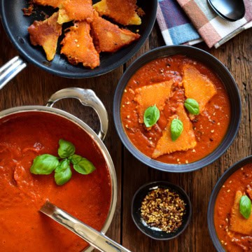 A pot of roasted red pepper tomato soup. Two bowls of soup served with three inside-out grilled cheese dumplings and fresh basil.
