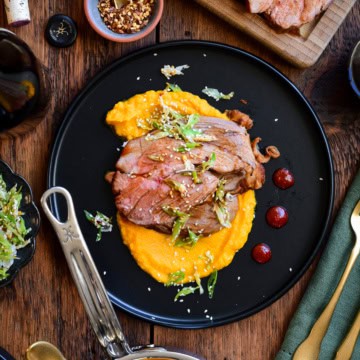 A plate with slices of gochujang honey glazed lamb on a bed of carrot puree. Surrounded by a pot of puree and more lamb slices.