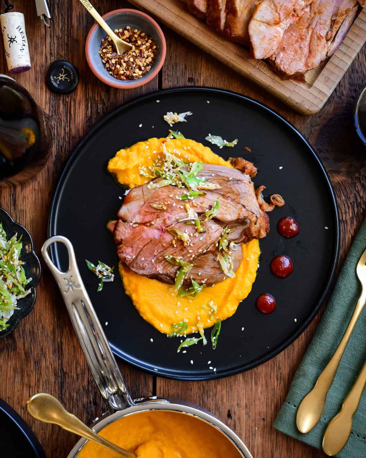 A plate with slices of gochujang honey glazed lamb on a bed of carrot puree. Surrounded by a pot of puree and more lamb slices.