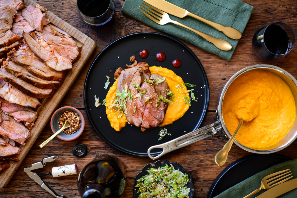 A plate with slices of gochujang honey glazed lamb on a bed of carrot puree. Surrounded by a pot of puree and more lamb slices.