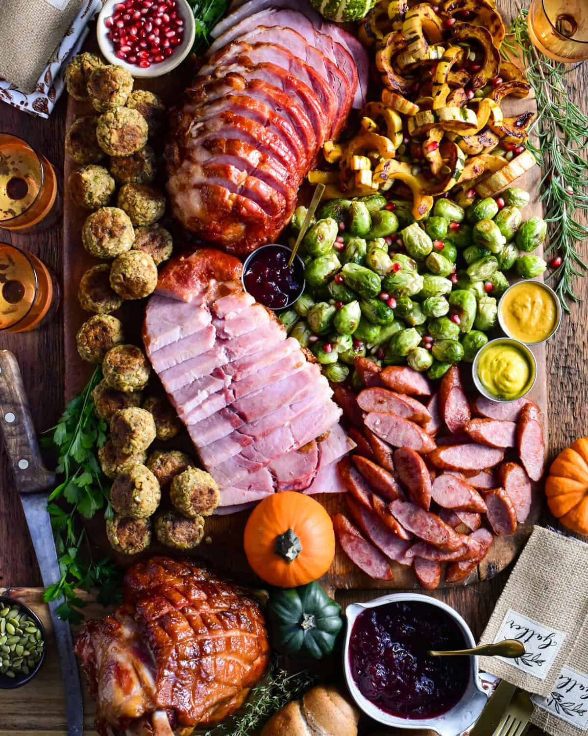 Slices of festive ham and turkey kolbassa are artfully arranged on a board with roasted Brussels sprouts, caramelized delicata squash, stuffing balls, cranberry sauce, and soft dinner rolls.