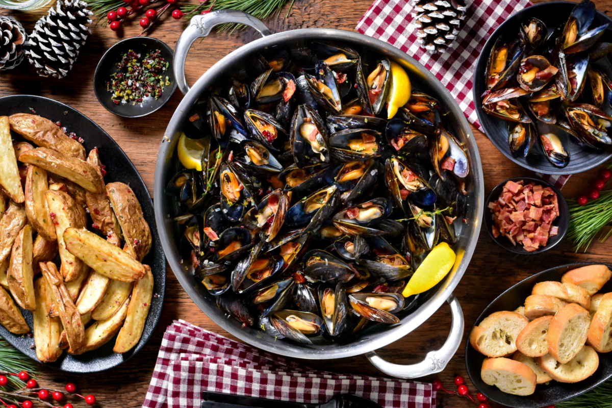 A pot of PEI Mussels in Cider Broth with Bacon and Cranberry Salt. they are served bread, lemon wedges and salt and malt vinegar potato wedges.